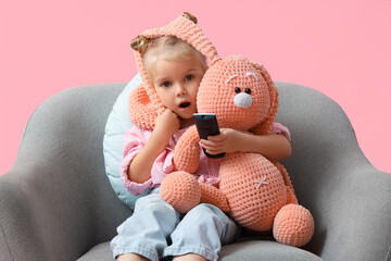 Shocked little girl with toy bunny in armchair watching TV on pink background