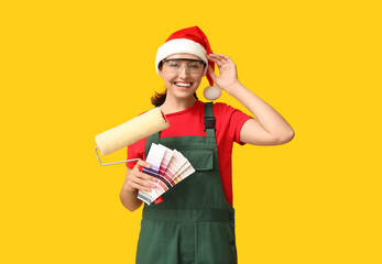 Beautiful female painter in Santa hat with roller and color palettes on yellow background