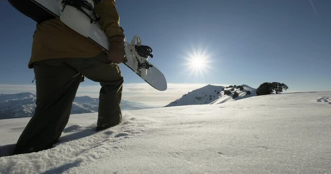 Close-up snowboarder walking in deep snow to the mountain peak, extreme off-trail freeriding adventure. A man with snowboard in hand walking through the pristine, knee-deep snow