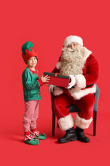 Santa Claus and cute little elf with Christmas gift on red background