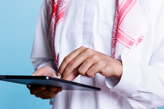 Muslim man wearing islamic traditional thobe making touch gestures with fingers on digital tablet screen closeup. Arab person using portable gadget doing zoom in on touchscreen