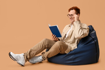 Young man sitting on beanbag and reading book against color background