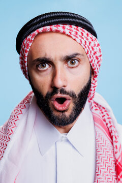 Muslim man with open mouth wearing traditional checkered ghutra headscarf studio closeup portrait. Arab person in islamic headdress showing emotion on face and looking at camera