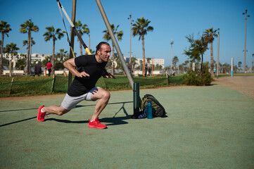 Full length portrait of determined Caucasian young adult sportsman doing lunges while working out with suspension straps