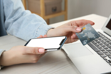 Online payment. Woman using credit card and smartphone with blank screen near laptop at white wooden table indoors, closeup