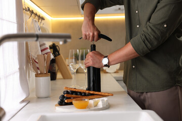 Romantic dinner. Man opening wine bottle with corkscrew at countertop in kitchen, closeup