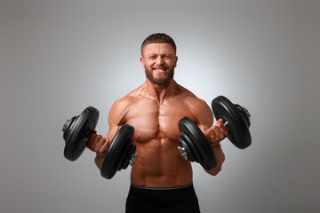 Fototapeta na wymiar Emotional young bodybuilder exercising with dumbbells on light grey background