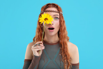 Surprised young hippie woman covering eye with sunflower on light blue background