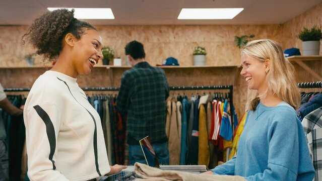 Female customer buying shirt from sales assistant at cash desk  and making contactless payment with credit or debit card in fashion or clothes store with customers in background - shot in slow motion