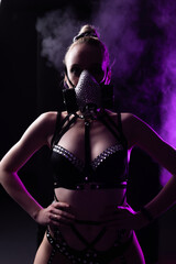 Portrait of a beautiful girl in black lingerie and leather harness. On the face is a respirator decorated with rhinestones. Contour purple light with smoke. Party concept, night club