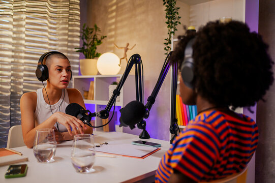 Serious multiracial girls are sitting in a home recording studio and filming a podcast while wearing headphones and discussing topics. - Powered by Adobe