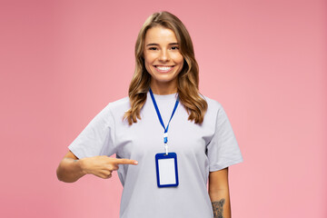 Portrait smiling attractive woman, seller consultant, manager wearing t shirt, pointing on blank badge looking  at camera isolated on pink background. Name card, Mockup 