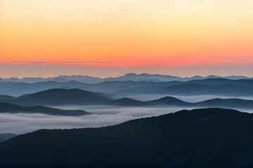 Fototapeta na wymiar 霧に覆われた山脈：オレンジと黄色のグラデーションが広がる空と、地平線上の薄いピンクの線、そして青と灰色の山々が霧やもやに覆われている、日の出または日没の風景