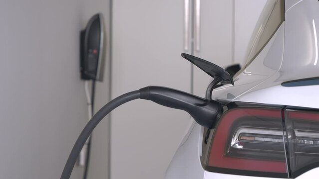 CLOSE UP: Electric vehicle connected to a charging station with a power cable. White electric car powering a rechargeable engine battery in home garage. Efficient alternative for modern transportation