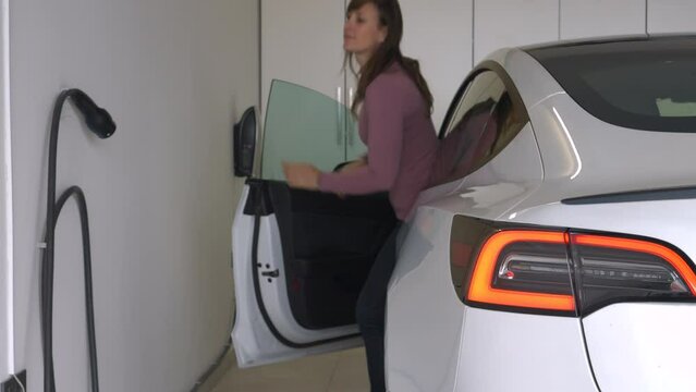 CLOSE UP: After parking in garage, a woman plugs in an electric car for charging. Young female driver connects black power cord to her white electric vehicle to recharge engine battery after driving.