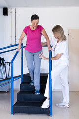 Vertical photo of a middle aged brunette woman in pink shirt. She is doing rehabilitation with her physiotherapist who is helping her.