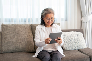 asian senior woman sitting on couch,relaxing in the living room,using internet online on tablet...