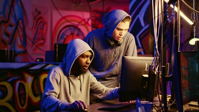 New member in hacking group receiving guidance from knowledgeable rogue programmer acting as his mentor in hidden base. African american hacker teaches script kiddie how to hack computers