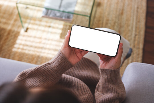Top view mockup image of a woman holding mobile phone with blank desktop white screen while sitting on a sofa at home