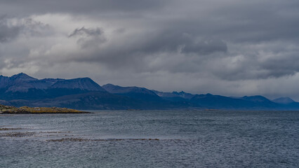 A picturesque mountain range of the Andes against a background of cloudy sky and ocean. An island with sparse vegetation is visible. Argentina. Beagle Channel. Tierra del Fuego Archipelago.