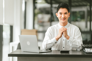 Attractive smiling male asian business person in white shirt working on laptop at desk with coffee cup and documents nearby.