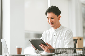 Fototapeta premium Attractive smiling male asian business person in white shirt working on laptop at desk with coffee cup and documents nearby.
