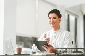 Attractive smiling male asian business person in white shirt working on laptop at desk with coffee cup and documents nearby.