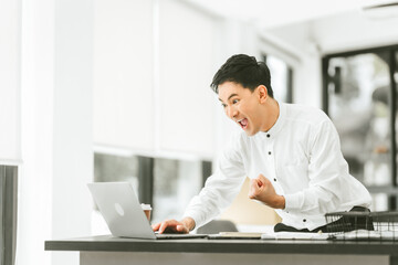Asian happy male business person in white shirt, fist raised in celebration, possibly excited or triumphant, sitting at desk with laptop.