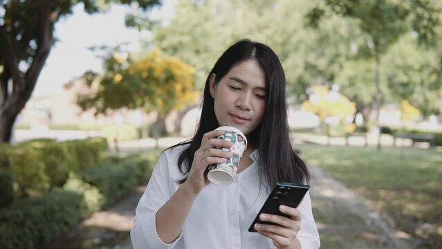 A young Asian woman is walking using a smartphone and drinking coffee in a takeaway cup in the park. Lifestyle and women concept.