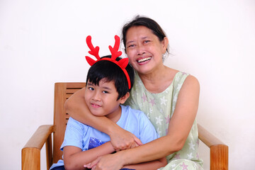 Asian mother hugging her chubby little boy wearing reindeer horn hair band, getting ready to...