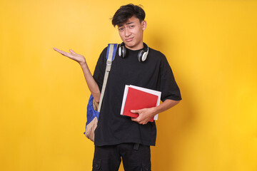 Young Asian student man wearing backpack and holding books, shrugging shoulders showing don't know...