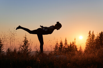 Young woman doing warrior 3 pose at fresh air. Silhouette of sporty female practicing yoga in...