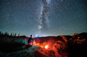 Man taking photos of starry sky with Milky way. Male photographer sitting on log and focusing...