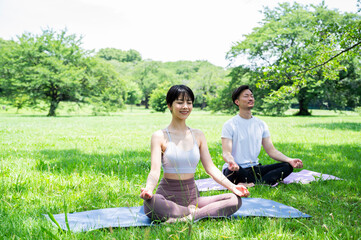 A beautiful Asian couple meditate on a sunny day, pulling out yoga mats in a park with lush green...