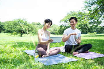 Happy Asian couple in sportswear sitting on the lawn looking at a digital tablet. Exercise, healthy living, urban living, environmental concerns, mental health, technology-enabled healthcare.