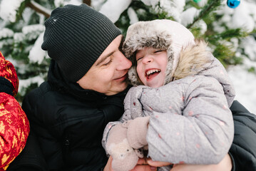 Obraz premium Dad hugging daughter sitting in snow in winter forest. Father embracing child girl near Christmas tree in park. Happy family walking snowy mountains, spending time together. Winter holidays. Closeup.