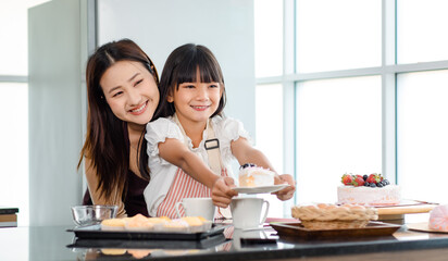 Asian pretty female baker pastry chef mother hugging little girl daughter wearing apron standing...