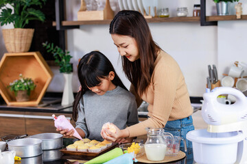 Asian female baker pastry chef mother helping teaching little girl daughter standing smiling holding piping decorating colorful whipped cream on cupcakes surface after baking tasty homemade bakery - obrazy, fototapety, plakaty