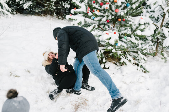 Man hold hands woman lying in snow near decorated Christmas tree in park. Couple smiling having fun in snowy winter forest. Young guy and girl in winter wear enjoying snowfall. Happy winter holidays.