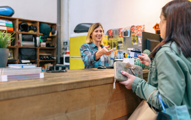 Female customer giving credit card to smiling blonde woman shop assistant to pay purchase on vintage store. Small business and local commerce concept. Selective focus on client. Left copy space. - obrazy, fototapety, plakaty