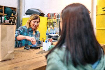 Smiling young female cashier passing credit card through store terminal to charge purchased items by customer in local shop. Happy shop assistant finishing payment of woman client in small business. - Powered by Adobe