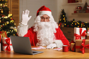 Santa Claus raising hand. Laptop, Christmas gifts and letters on table