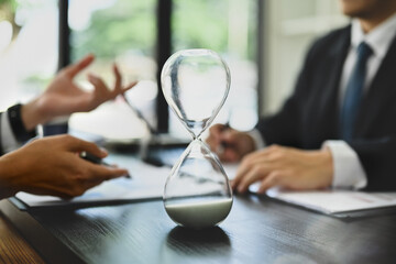Closeup hourglass on meeting table with blurred two businesspeople working on background - obrazy, fototapety, plakaty
