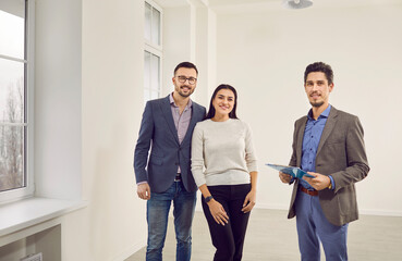 Portrait of young happy couple with realtor looking at camera and smiling. Professional real estate agent shows bright new apartment. Family ready to become homeowners. Real estate market concept.