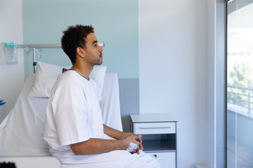 Profile of biracial male patient sitting on bed with iv drip in sunny hospital room