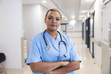 Portrait of happy biracial female doctor wearing scrubs and stethoscope in corridor at hospital
