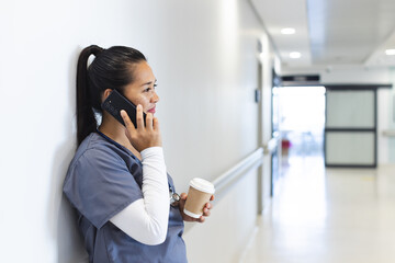 Fototapeta premium Biracial female doctor talking on smartphone, holding takeaway coffee in hospital, copy space