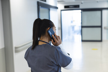 Biracial female doctor wearing scrubs talking on smartphone in hospital corridor