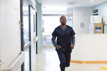 African american male doctor wearing scrubs running through corridor in hospital, copy space