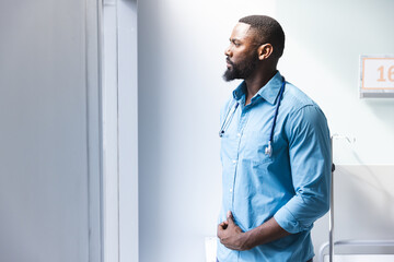 Fototapeta premium Thoughtful african american male doctor looking through window in hospital room, copy space